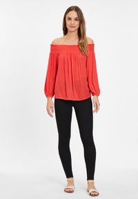 Blusa roja de un solo hombro con mangas largas, con textura fruncida en el escote, combinada con leggings negros y sandalias ligeras.