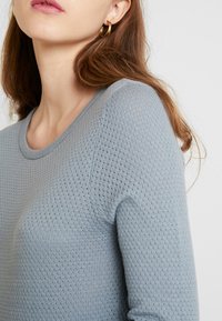 Helles blaues Strickpullover mit strukturiertem Muster, runden Ausschnitt, figurbetonten Ärmeln und glatter Oberfläche; getragen mit einem goldenen Creolenohrring.
