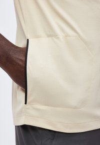 Main à la peau foncée dans la poche latérale d'une chemise texturée beige clair avec bordure noire, portée sur un pantalon gris foncé.
