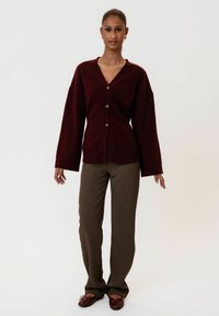 Bordo oversized kardigan s V-izrezom, s pet dugmadi i produženim rukavima, u kombinaciji s smeškastim ravnim hlačama i tamnim cipelama.