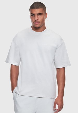 Young man with short hair and earring, wearing oversized white t-shirt and white pants, standing against plain gray background.