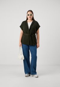 Tamsiai žalia siuvinėta apsiaustė trumpomis rankovėmis, surišta ties liemeniu, derinama su mėlynomis plačiomis kelnėmis ir maža balta rankine.