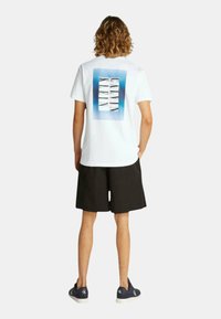 Camiseta blanca de algodón con un diseño rectangular en acuarela azul y el texto "Calvin Klein" en la parte posterior, combinada con pantalones cortos negros y zapatillas náuticas.