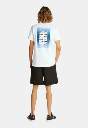 Camiseta blanca de algodón con un diseño rectangular en acuarela azul y el texto "Calvin Klein" en la parte posterior, combinada con pantalones cortos negros y zapatillas náuticas.