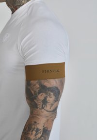 White fitted t-shirt with a brown elastic arm band labeled "SIKSILK." Visible detailed tattoos on the arm.