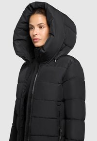 Sort dunjakke med en overdimensioneret hætte, lynlås foran og elastiske manchetter. Har en firkantet quiltedesign; stoffet ser glat og mat ud.