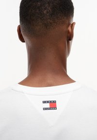 Set bagfra af en person iført en hvid skjorte med et Tommy Hilfiger-logo nær halsudskæringen, på en ensfarvet hvid baggrund.