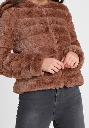 Ruskea pehmeä tekoturkistakki, jossa vaakasuorat tikatut osiot, ylle puettuna tummanharmaiden farkkujen kanssa, kädet kevyesti ristissä vyötäröllä.