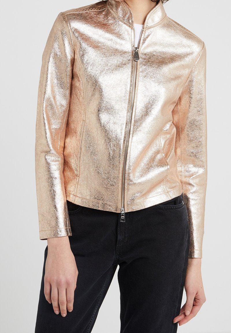 Metallisch roségoldene Kunstlederjacke mit hohem Kragen und Reißverschluss. Sie hat lange Ärmel und eine glatte Textur. Stylisch mit schwarzen Hosen kombiniert.