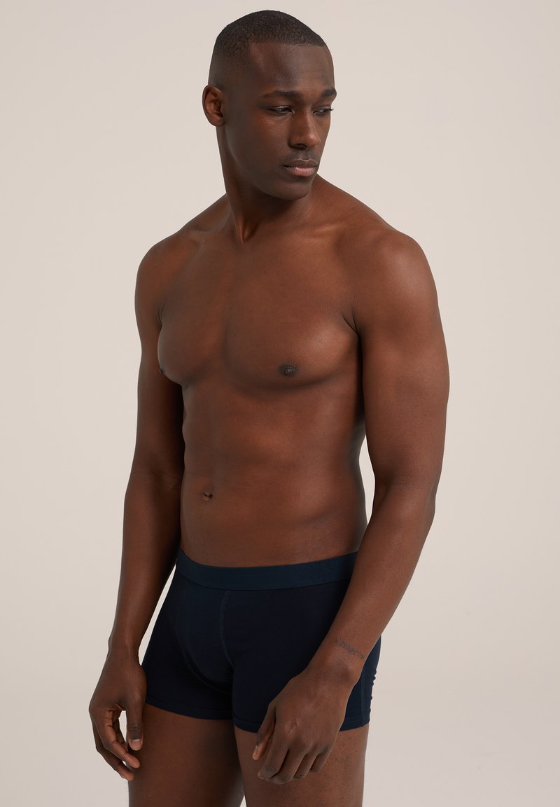 Morsko modri bokserji z mehkimi pasom, prilegajoča zasnova in gladka tkanina, nosi jih brezmajhen moški model z atletskim telesom.