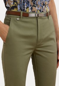 Close-up of olive green pants with side pockets, a brown leather belt with silver buckle, and a multicolored patterned tucked-in shirt.