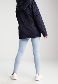 Osoba v námořnickém mikině s kapucí, světle modrých skinny džínách a bílých teniskách stojící zády k objektivu na jednoduchém světlém pozadí.