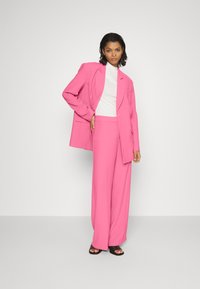 Fato rosa ajustado com um blazer estruturado e calças de perna larga, combinado com um suéter de gola alta branco e sandálias pretas de dedos à mostra.