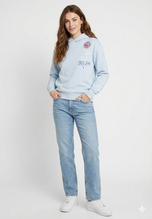 Junge Frau mit langen braunen Haaren, die einen hellblauen Kapuzenpullover mit Schriftzug und hellblaue Jeans trägt, mit den Händen in den Taschen stehend und lächelnd.