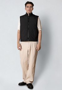 Hombre con chaleco sin mangas negro con cremallera sobre una camiseta beige de manga corta, pantalones anchos beige y zapatos negros, de pie contra un fondo liso.