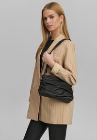 Borsa a tracolla nera in pelle con design arricciato, texture liscia e una sola tracolla, esposta su uno sfondo grigio uniforme.