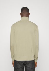 Sudadera verde oliva claro con cuello con cremallera, corte relajado y detalles de costura texturizada en los hombros y en los puños. Combinada con jeans oscuros.