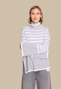 Gråstripete strikkegenser med høy krage, med bredere ermer og løs passform. Synlig tekstur kontrasterer med glatte hvite detaljer.