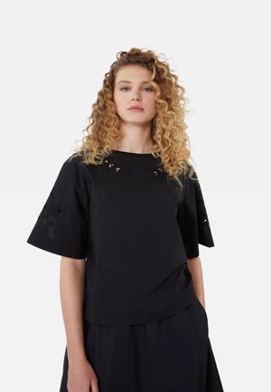 Donna con capelli biondi ricci che indossa una maglia nera larga con motivi floreali ritagliati sulle maniche e sulla parte superiore del petto su uno sfondo semplice.