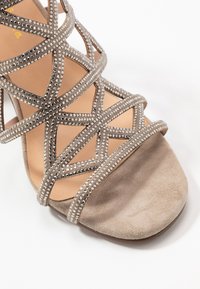 Sandalo con tacco in suede beige con cinturini incrociati decorati con gemme trasparenti, design open toe e suola interna imbottita.