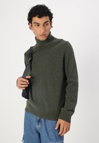Pull à col roulé vert foncé en tricot doux, avec des poignets et un ourlet côtelés. Porté avec un sac à épaule noir réglable.