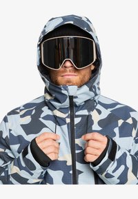 Blå og grå camouflage snejakke med høj krave, lynlås lukning og justerbar hætte. Har sorte håndledsmanchetter og et mønstret tekstur.