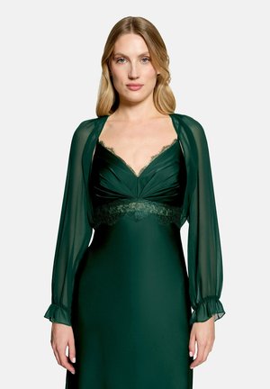 Femme aux cheveux blonds portant une robe vert foncé avec des manches longues transparentes et des détails en dentelle au niveau de l'encolure et de la taille, debout devant un fond clair.