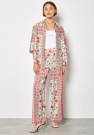 Kobieta ubrana w kolorowy garnitur w kwiatowy wzór z szerokimi spodniami i otwartą marynarką na białym topie oraz białych sandałach na obcasie.