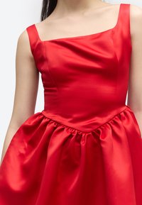 Közelről látható egy személy, aki ujjatlan piros szatén ruhát visel, négyzet alakú kivágással és gyűrt derékrésszel.