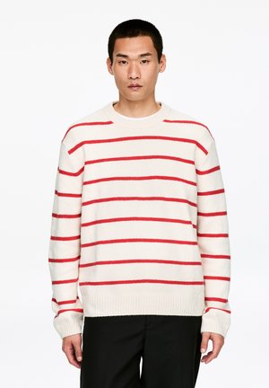 Pull ras du cou en maille crème avec de larges rayures horizontales rouges. Comprend des poignets et un ourlet côtelés, une texture douce, une coupe décontractée. Pantalon noir porté.
