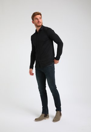 Camisa negra de manga larga con botones en el frente, combinada con jeans oscuros y botas de tobillo beige. Tejido suave y elástico, con un ajuste entallado.