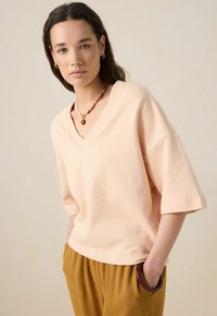 Haut rose clair à col en V avec des manches courtes, en textile doux, coupe décontractée. Porté avec un pantalon marron et un collier en perles avec un pendentif.