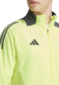 Veste de sport jaune vif avec un col noir et des accents noirs, dotée d'une fermeture éclair et de trois bandes noires sur les épaules.
