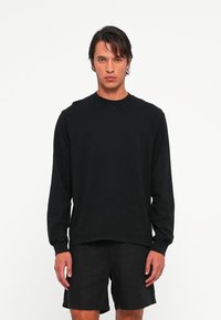 Man met halflang donker haar, gekleed in een eenvoudige zwarte longsleeve en zwarte shorts, staand tegen een witte achtergrond.