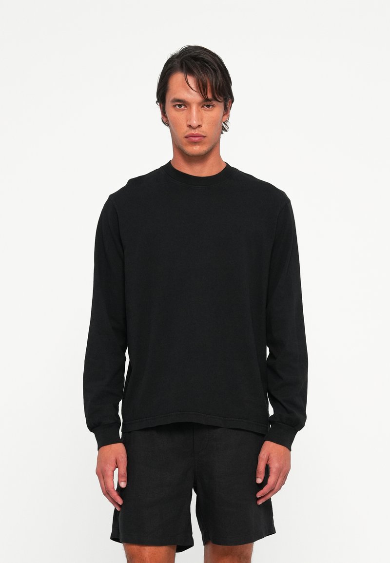 Man met halflang donker haar, gekleed in een eenvoudige zwarte longsleeve en zwarte shorts, staand tegen een witte achtergrond.