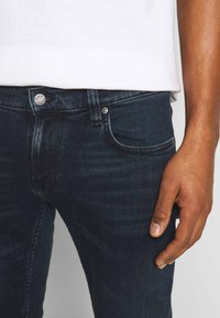 Mörkblå denimjeans med slim fit, med dragkedja, framficka och metallknappdetalj i midjan. Slät textur.