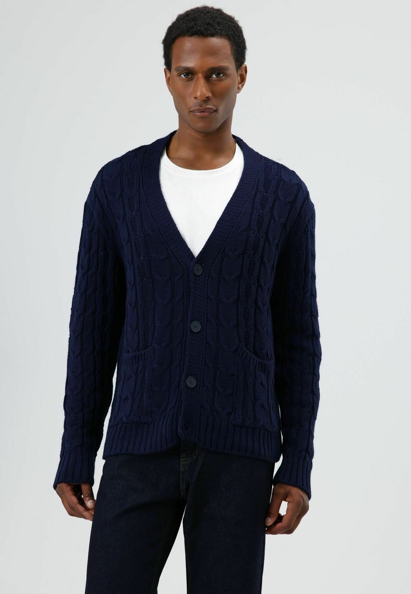 Marineblauer Strickcardigan mit V-Ausschnitt, Knopfverschluss und zwei Vorderseitentaschen. Strukturierter Stoff und gerippte Bündchen verleihen dem Design mehr Tiefe.