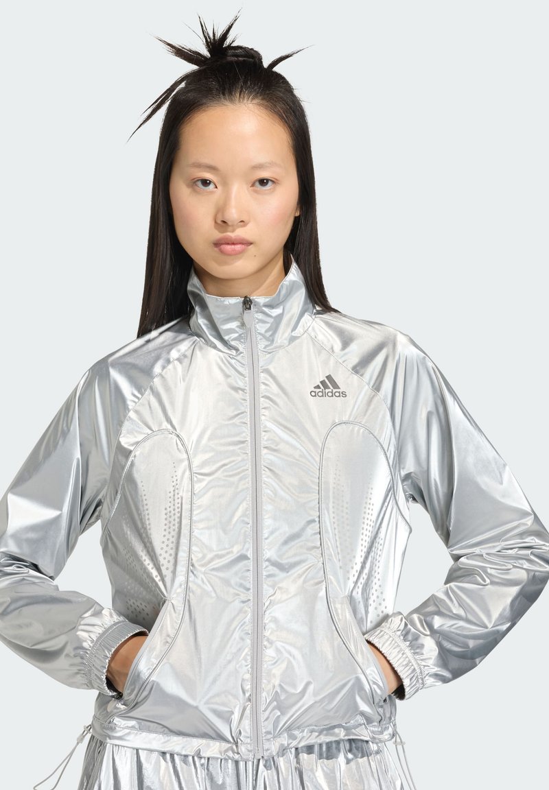 Veste de sport argentée avec col montant, fermeture éclair à l'avant et poignets élastiques. Caractérisée par des détails perforés et un matériau doux, brillant et léger.