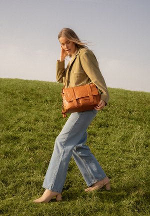 Woman walking on grassy hill wearing beige jacket, light blue jeans, nude heels, and carrying a large brown leather shoulder bag.