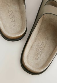 Bruna och svarta sandaler med en slät läderoverdel, vadderad mocka fotbädd och texturerad gummisula. Har ”NEXT” präglat på fotbädden.