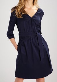 Námořnická modrá zavinovací šaty vyrobené z hladké látky, s výstřihem do V, tříčtvrtečními rukávy, opaskem v pase a rozšířenou sukní.