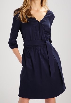 Granatowa sukienka typu wrap, wykonana z gładkiego materiału, z dekoltem w kształcie V, trzyczwartowymi rękawami, paskiem w talii i rozkloszowanym dołem.