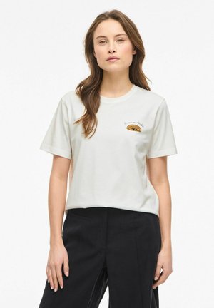 Femme aux longs cheveux bruns portant un T-shirt blanc avec un petit logo et un motif texte sur la poitrine, associée à un pantalon noir, immobile.