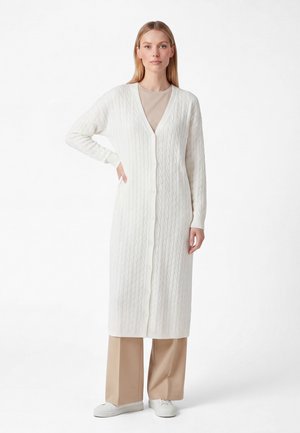 Kvinde iført en lang hvid kabelstrikket cardigan med knapper over beige vidde bukser og hvide sneakers, stående mod en ensfarvet baggrund.