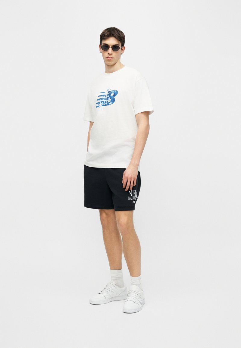 Jeune homme portant des lunettes de soleil, un t-shirt blanc New Balance avec logo bleu, un short noir avec texte blanc New Balance, des chaussettes blanches et des baskets blanches.