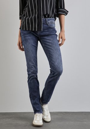 Jean skinny bleu foncé en denim, avec un design classique à cinq poches, porté avec des baskets blanches. Le haut est noir avec des rayures blanches.