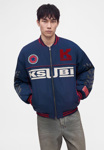 Jeune homme portant une veste zippée bleu marine avec des patchs rouges et blancs, debout avec les mains dans les poches sur un fond blanc.