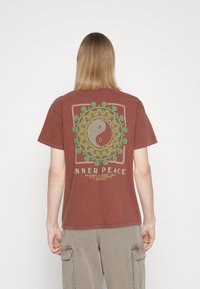 Brun bomull t-shirt med en stor grafisk design som visar en yin-yang symbol och mandalamönster, kompletterat med gröna och vita accenter.