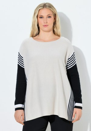 Mujer joven con cabello rubio largo, vestida con un suéter suelto a rayas blancas y negras y pantalones negros, de pie frente a un fondo blanco.