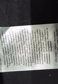 Pflegeetikett auf Stoff, rechteckig, strukturiertes weißes Material mit schwarzem Text, der die Zusammensetzung des Stoffes und Pflegeanweisungen in mehreren Sprachen auflistet.